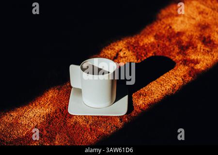 A white coffee cup on a saucer casts a long shadow across a textured brown soft carpet, contrasting with the bright light. Morning drink at cozy home. Stock Photo