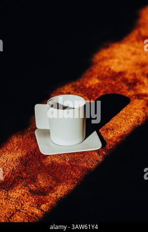 A white coffee cup on a saucer casts a long shadow across a textured brown soft carpet, contrasting with the bright light. Morning drink at cozy home. Stock Photo
