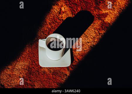 A white coffee cup on a saucer casts a long shadow across a textured brown soft carpet, contrasting with the bright light. Morning drink at cozy home. Stock Photo