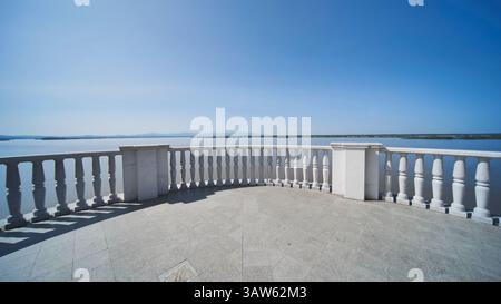 Marble terrace with ornate balusters framing serene lake waters, reflecting cloudless sky and radiating tranquil landscape atmosphere in Khabarovsk. Stock Photo