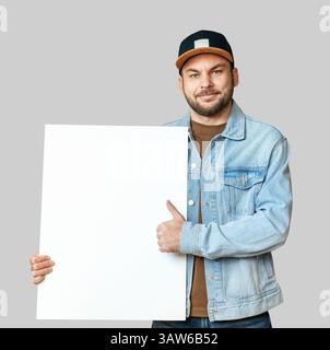 mockup isolated young adult man in denim jacket and cap holding white empty board and gesturing thumbs-up Stock Photo