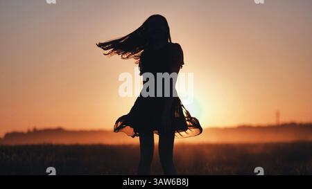 Carefree woman silhouetted against sunset sky, dancing wildly in grassy field, flowing hair capturing joy, freedom, and natural connection Stock Photo