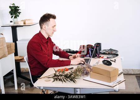 Independent jewelry maker managing his online store using a laptop at a workbench filled with tools and eco packaging. Creative entrepreneurship Stock Photo