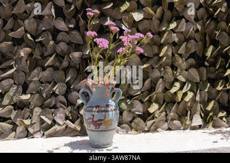 A painted ceramic vase with purple blossoms sits in the sun against a leafy backdrop on a Greek island. Stock Photo