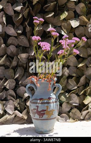 A painted ceramic vase with purple blossoms sits in the sun against a leafy backdrop on a Greek island. Stock Photo
