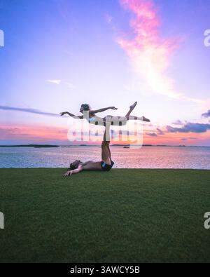 Sporty couple man and woman silhouettes doing an acrobatic yoga pose on grass in front of beautiful sunset over the sea. Stock Photo