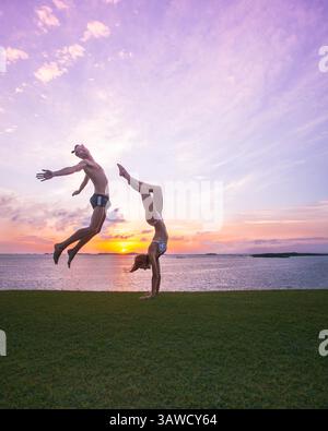 Sporty couple man and woman silhouettes doing an acrobatic yoga poses on grass in front of beautiful sunset over the sea. Male is doing dancer jump. Stock Photo