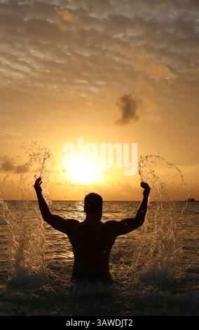 Silhouette of a man playfully splashing water as the sun sets over the ocean, creating a stunning backdrop of warm colors and dramatic clouds. Stock Photo