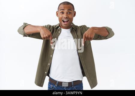 Portrait of excited handsome african-american man talking about big news, great discounts, pointing fingers down, look happy camera beaming smile from Stock Photo