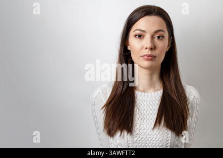 Close-up of a calm, serious, emotionless young woman with brown hair, isolated on a white background. Stock Photo
