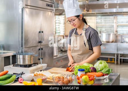 Young female chef wearing a chef hat and striped apron  is smiling and cutting fresh tomato. Fresh vegetables and cooking ingredients are laid out on Stock Photo