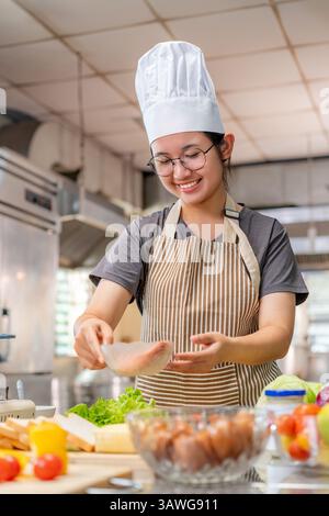 A young female chef wearing a striped apron and chef hat preparing fresh vegetables and other ingredients in a commercial kitchen. She is focused whil Stock Photo