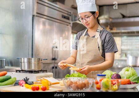A cheerful young female chef wearing a striped apron and white chef hat is seen preparing a fresh sandwich in a commercial kitchen. Surrounded by fres Stock Photo
