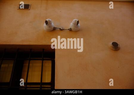 Cremona, Italy March 15th 2025 Two white security cameras are mounted on a peach colored wall, connected by a cable, above a barred window, enhancing Stock Photo