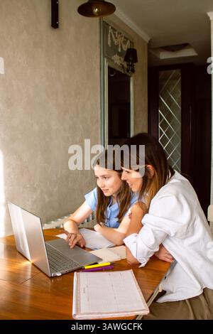 Happy mother with preteen daughter using laptop in home kitchen. Working on school project. Helping with homework using technologies Stock Photo