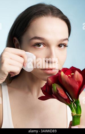 Woman using gua sha tool while holding red flowers against blue background Stock Photo