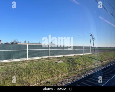 A rural railway landscape with a metal noise barrier fence running parallel to the train tracks. The scene includes green grass, power lines, and a cl Stock Photo