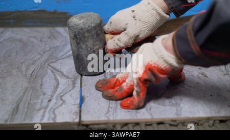 Tiler placing ceramic tiles on bathroom floor, using rubber mallet and leveling system during renovation work Stock Photo