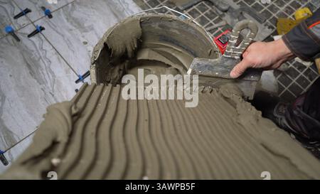 Construction worker spreading tile adhesive on floor using notched trowel, preparing for tile installation Stock Photo