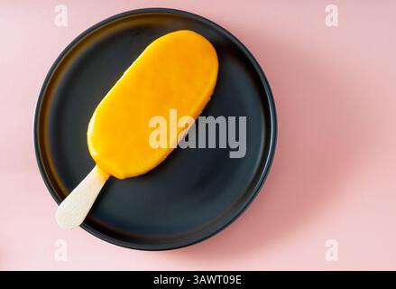 A top-down shot shows a vibrant orange popsicle lying on a dark blue or black round plate. The background is a soft, light pink color. Stock Photo