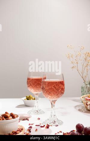 Two glasses of rose wine on a white tablecloth. On the table there are a small bowl with peanuts, red grapes, a bread basket and a green vase. Stock Photo