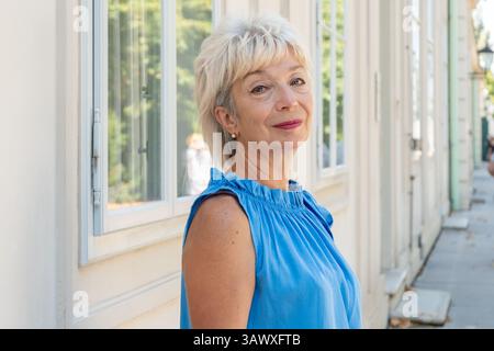 A stylish woman aged 65-70 in a blue outfit stands outside a building with window. Stock Photo
