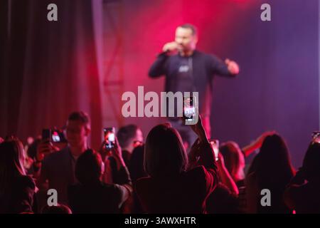 Crowd enjoying live music performance, taking photos with glowing red stage lights in background. Stock Photo