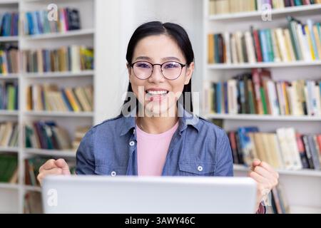 An excited asian woman wears glasses and looks at a laptop with an open mouth in a library, celebrating. Bookshelves fill the background. Stock Photo