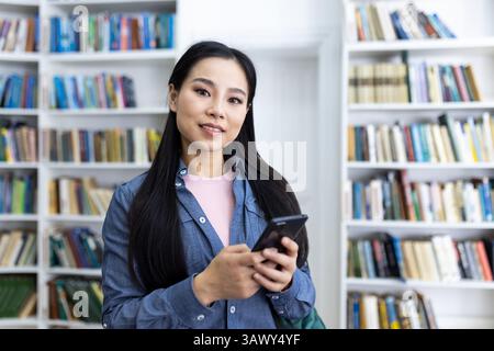 A smiling asian woman holding a smartphone in a library setting, surrounded by books and shelves. She is likely a student or a reader. Stock Photo