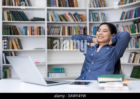 A relaxed woman smiles with her eyes closed, enjoying a break in the library. Her arms rest behind her head as she sits at her desk. Bookshelves fill the background. Stock Photo