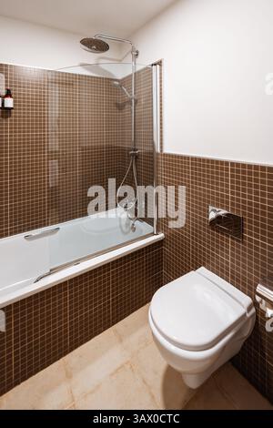 Modern bathroom with wall-mounted toilet, white bathtub with overhead rain shower and handheld shower, brown mosaic wall tiles, glass partition, ceram Stock Photo