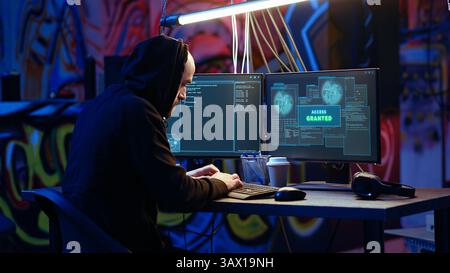 Hooded hacker in graffiti painted hideaway targeting vulnerable unpatched connections, having burst of joy after getting access granted past security and stealing valuable sensitive data Stock Photo