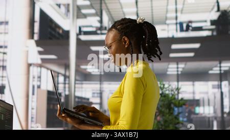 Computer scientist working, doing software debugging using notebook, troubleshooting errors. Woman standing in office, fixing coding issues using laptop, solving tasks, camera A Stock Photo