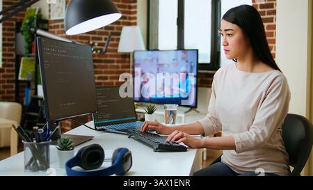 Professional software developer types code on a laptop in a sleek workspace. Asian female freelancer doing remote work on app development, software engineering and agile coding practices. Camera A. Stock Photo