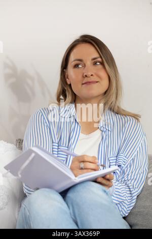 Woman with thoughtful expression fills diary sitting on sofa Stock Photo