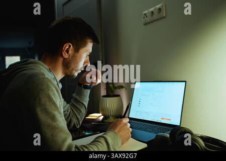 Concentrated freelancer working late at home office. Focused man in hoodie writes notes while working on laptop. Concept of night work Stock Photo