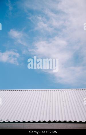 Crisp, white corrugated rooftop against a bright blue sky with soft, fluffy clouds. A minimalist view showcasing architectural detail. Stock Photo