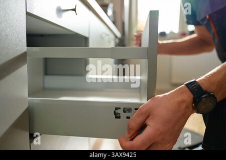 Man assembling kitchen drawer in modern cabinet using screwdriver. Handyman installing and adjusting drawer slide mechanism in contemporary kitchen. F Stock Photo