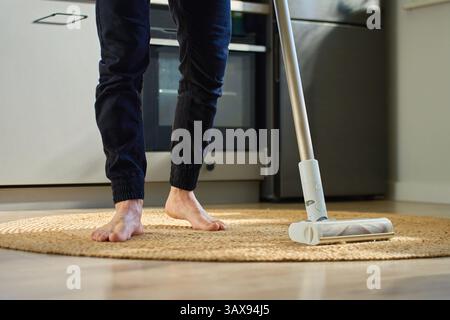 Barefoot man vacuuming round woven rug on wooden floor in kitchen. Person uses handheld stick vacuum cleaner in apartment. Concept of daily home clean Stock Photo