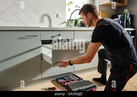 Man assembling kitchen drawer in modern cabinet using screwdriver. Handyman installing and adjusting drawer slide mechanism in contemporary kitchen. F Stock Photo