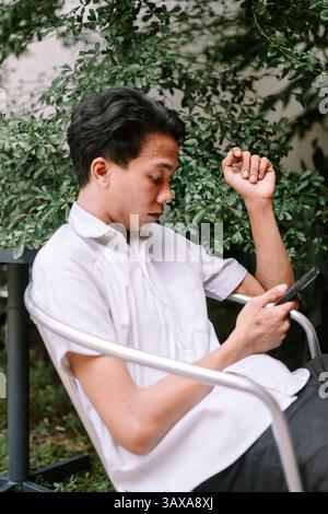 A relaxed young man is using his smartphone, enjoying a calm moment outdoors surrounded by lush greenery. He seems thoughtfully engaged with the conte Stock Photo