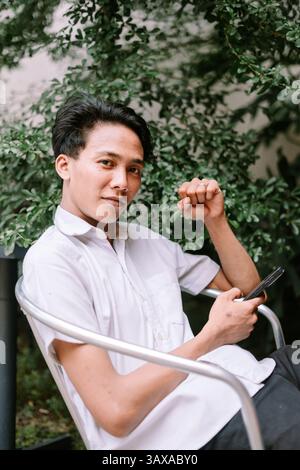 A young asian man casually rests outdoors, using his smartphone amidst a backdrop of vibrant green foliage. He appears relaxed and connected. Stock Photo