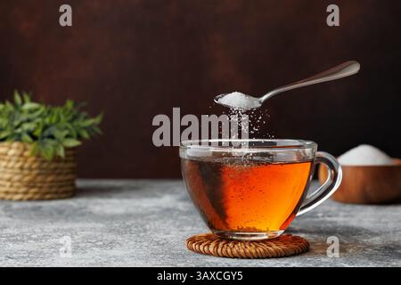 White sugar falling from teaspoon into glass cup of hot tea placed on coaster against dark brown background Stock Photo
