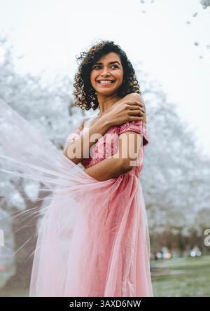 A woman smiles wrapping her arms around herself in a long pink dress Stock Photo