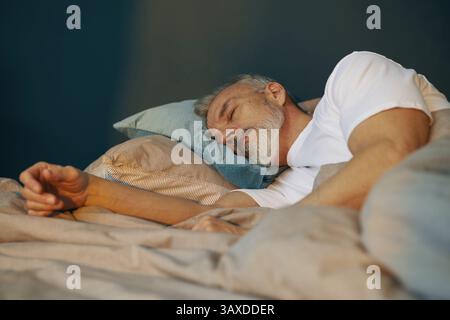 Elderly man lying on his side on comfortable bed, resting his head on pillows and sleeping peacefully with relaxed expression Stock Photo