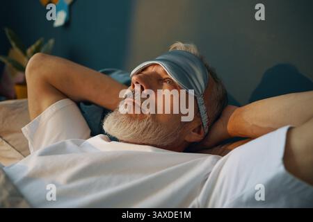 Senior man reclining on bed, wearing sleep mask and looking serene. Background shows minimalistic decor with plants and relaxed atmosphere Stock Photo