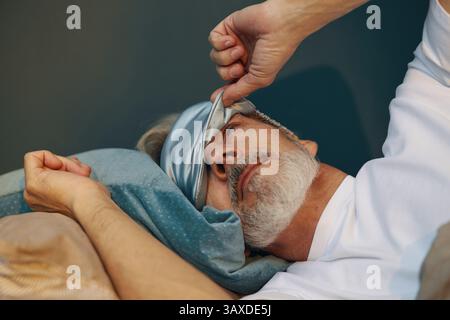Caucasian senior man lying on bed adjusting sleep mask while tucking in comfortably with head resting against soft pillow Stock Photo