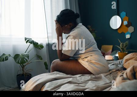 African American woman sitting on bed with hand on face, surrounded by potted plants and decor. Natural morning light entering through window creating Stock Photo