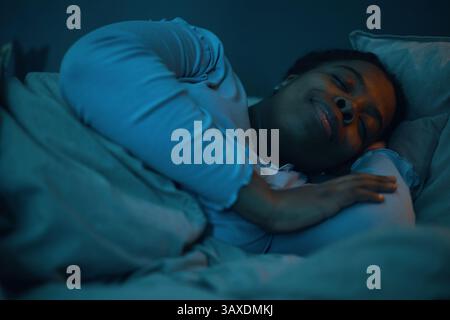 African American woman resting comfortably on bed, smiling with eyes closed while snuggled into pillow. Soft lighting giving a calm and tranquil ambia Stock Photo