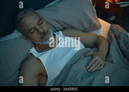 Elderly man peacefully resting with head on pillow while sleeping in bed under a cozy blanket holding onto the bedding Stock Photo